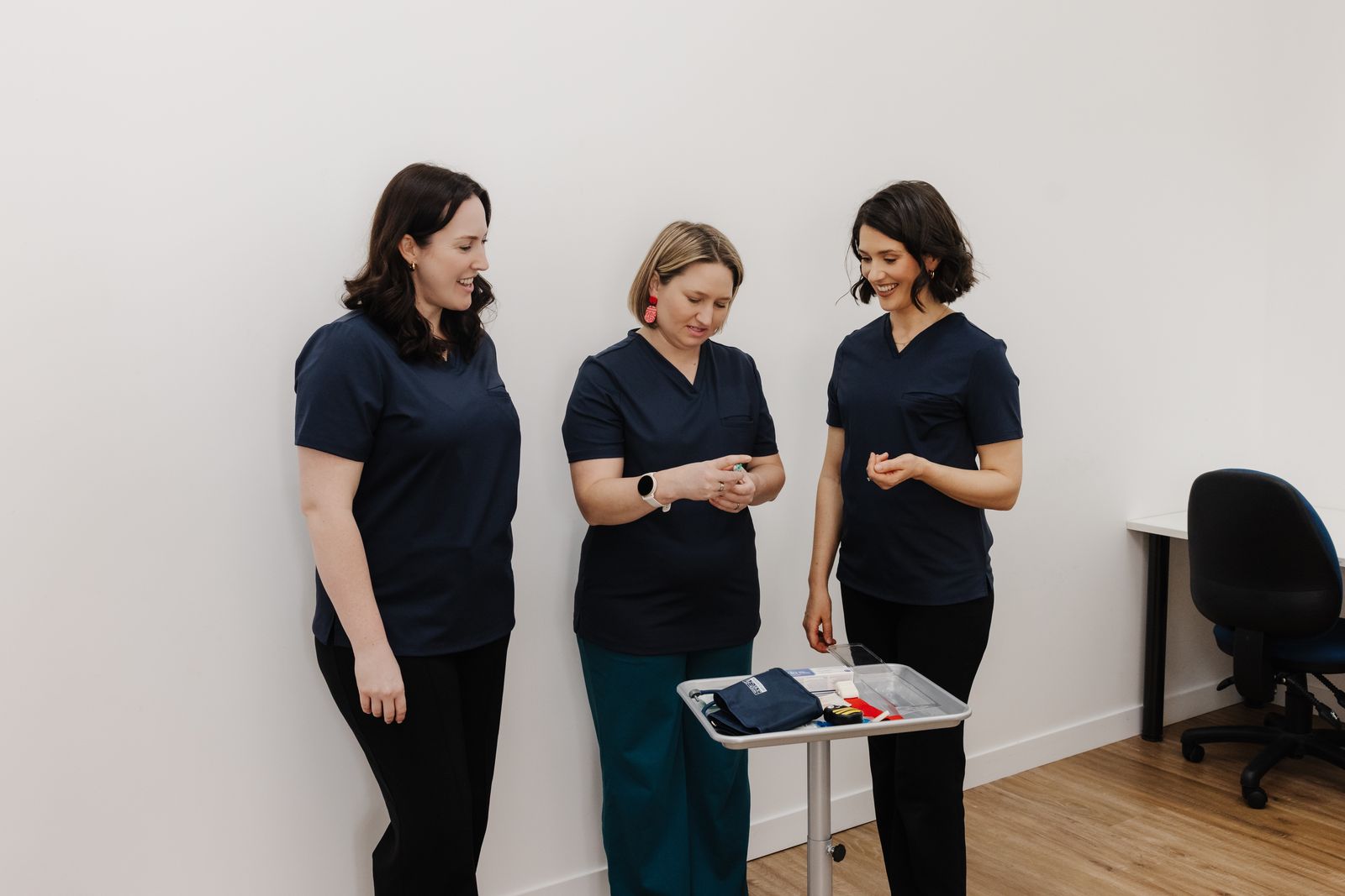 Three smiling Brave Steps Health clinicians in navy scrubs looking at healthcare practice equipment on a clinic trolley